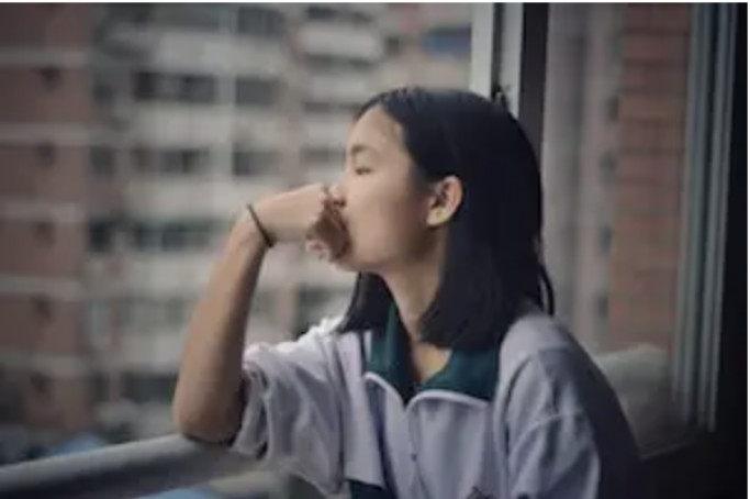 A young person in a gray hoodie leans on a windowsill, looking out at apartment buildings.
